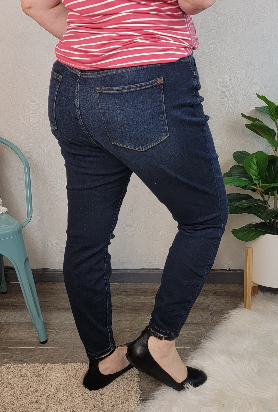Wearing Judy Blue's Mandy Skinny Jeans, a red and white striped top, and black flats, a person stands on a wooden floor beside a green chair and potted plant. The photo is taken from behind, focusing on the lower half.