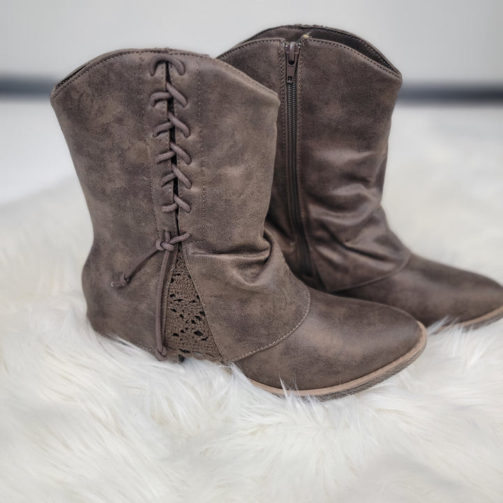 The Sassy Booties By Very G are taupe faux suede boots by Very G, featuring side zippers, decorative laces, and crochet edge details, shown on a white fluffy surface.