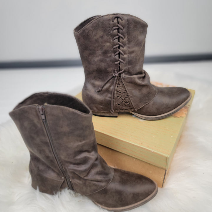 The Sassy Booties by Very G are brown, mid-calf lace-up booties with side zippers and decorative laces. One stands on a shoebox, the other beside it on a white fuzzy surface. Brand: Very G.
