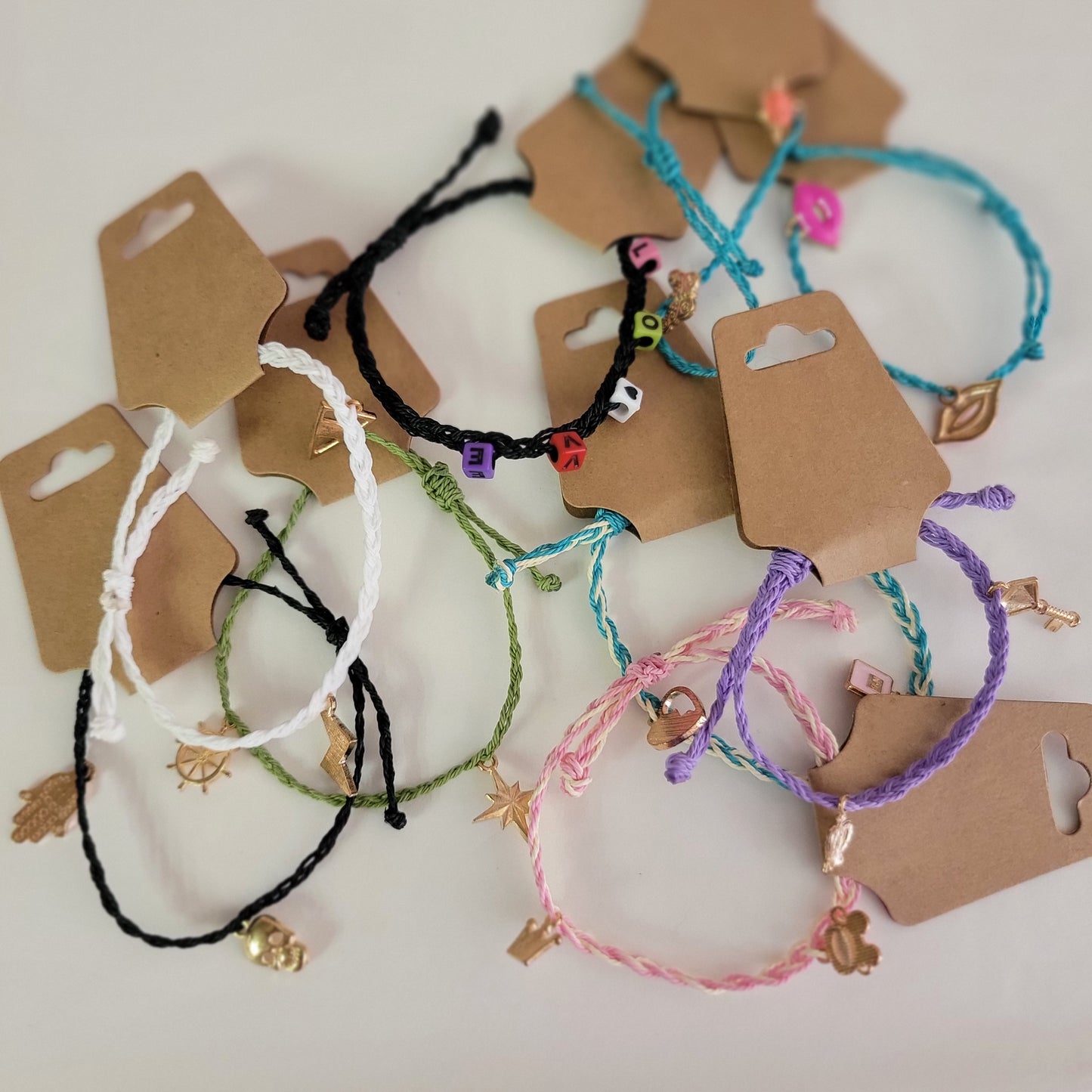Make By Molly: Charm Bracelets by Molly Thurmer—colorful braided string bracelets with assorted charms and bead letters—are displayed on a white surface, each attached to a brown paper retail tag.
