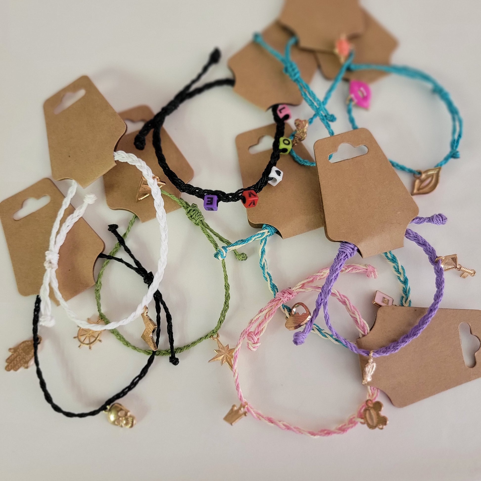 Make By Molly: Charm Bracelets by Molly Thurmer—colorful braided string bracelets with assorted charms and bead letters—are displayed on a white surface, each attached to a brown paper retail tag.