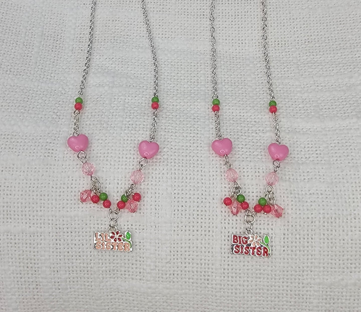 Two Girls Charm Jewelry necklaces by 2HOPE Accessories, Inc—one "Big Sister," one "Little Sister"—feature pink, green, and red beads with a pink heart and charm. Displayed on white fabric, they're perfect as necklace sets for sisters!.