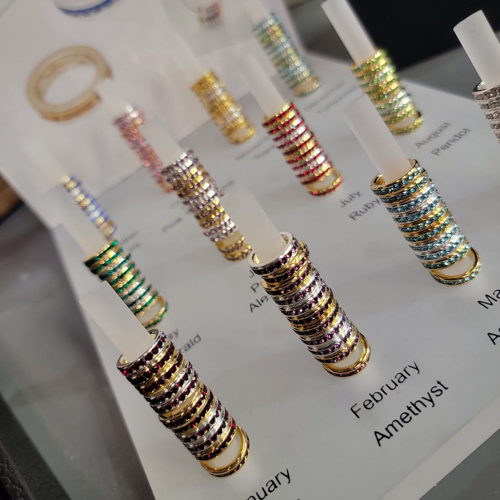 Rows of colorful Birthstone Eternity Charms by S&A Fashions are arranged on white rods, each labeled by month and gemstone—like Amethyst for February—and displayed with matching silver or gold pendant options in the case.