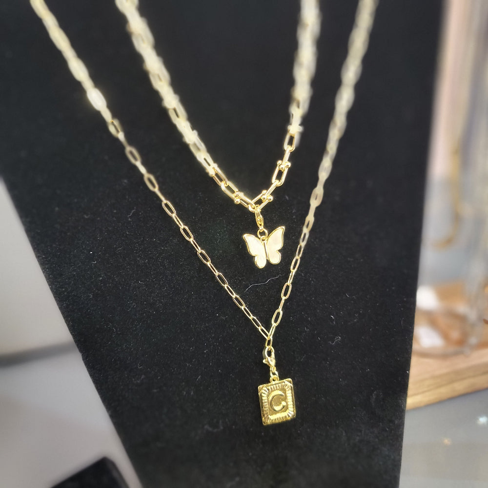 Three gold chains on a black stand: one with a butterfly pendant, one with a rectangular locket with the letter C, and the S&A Fashions Paperclip Chain with a simple link design.