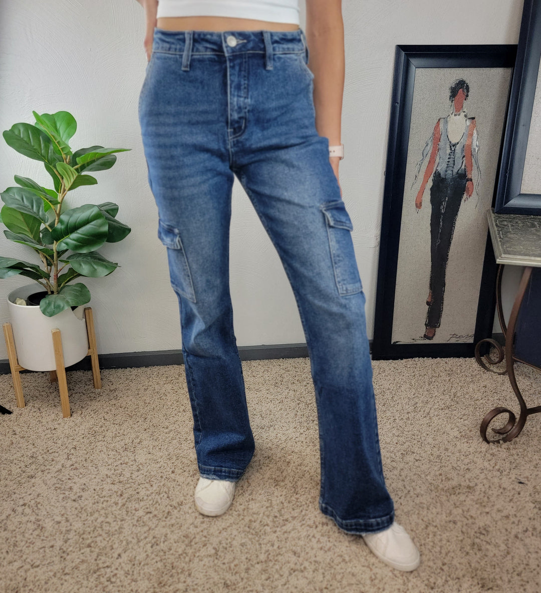 A person wearing KanCan's Holly Wide Leg Flare jeans, a white crop top, and white shoes stands on a carpeted floor with a potted plant and a framed fashion illustration in the background.