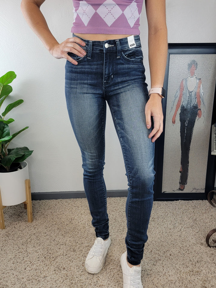 Wearing Judy Blue's Constance Skinny Jeans - Tall! in dark blue with a 34" inseam, a sleeveless purple argyle top, and white sneakers, a person stands on beige carpet near a framed fashion illustration and potted plant, hand on hip.