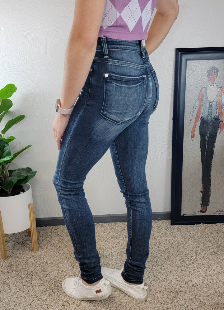 A person wearing dark blue Constance Skinny Jeans - Tall! by Judy Blue, a purple sleeveless argyle top, and white sneakers stands on beige carpet near a potted plant and framed artwork of a figure in jeans.