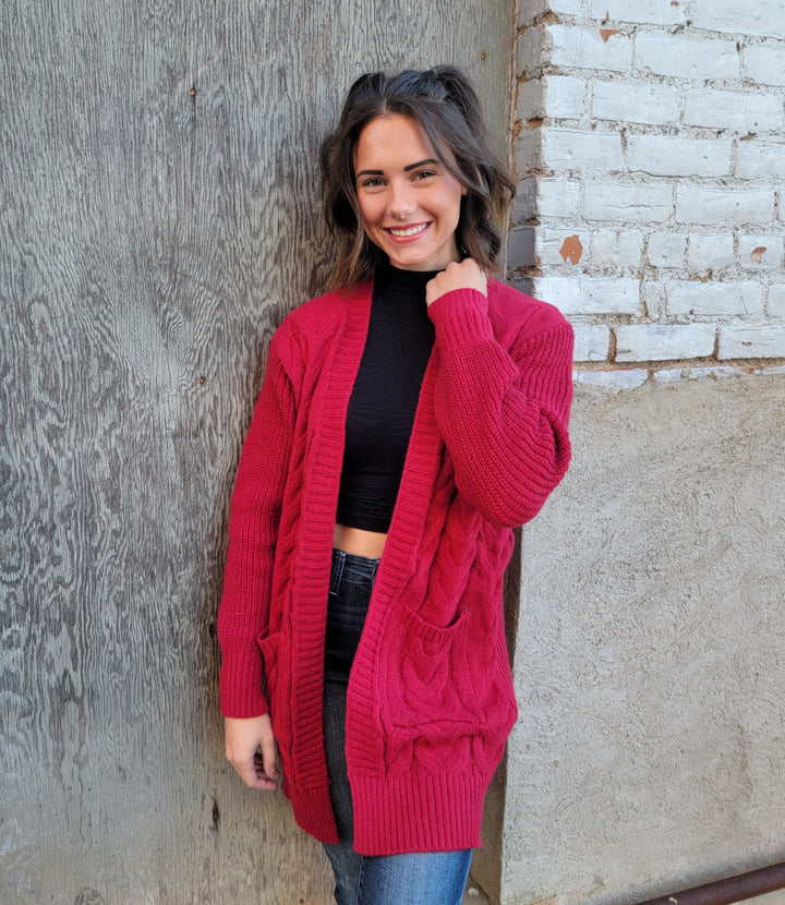 A woman with shoulder-length brown hair, in a black crop top, blue jeans, and a red Nali Elaine Cardigan, smiles against a wall with wood and white brick accents.