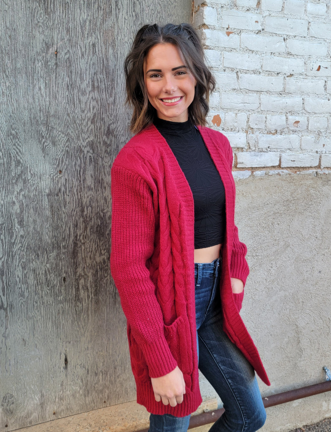 A young woman with shoulder-length dark hair smiles outdoors by a rustic wall, wearing the Nali Elaine Cardigan in red over a black turtleneck crop top and blue jeans.