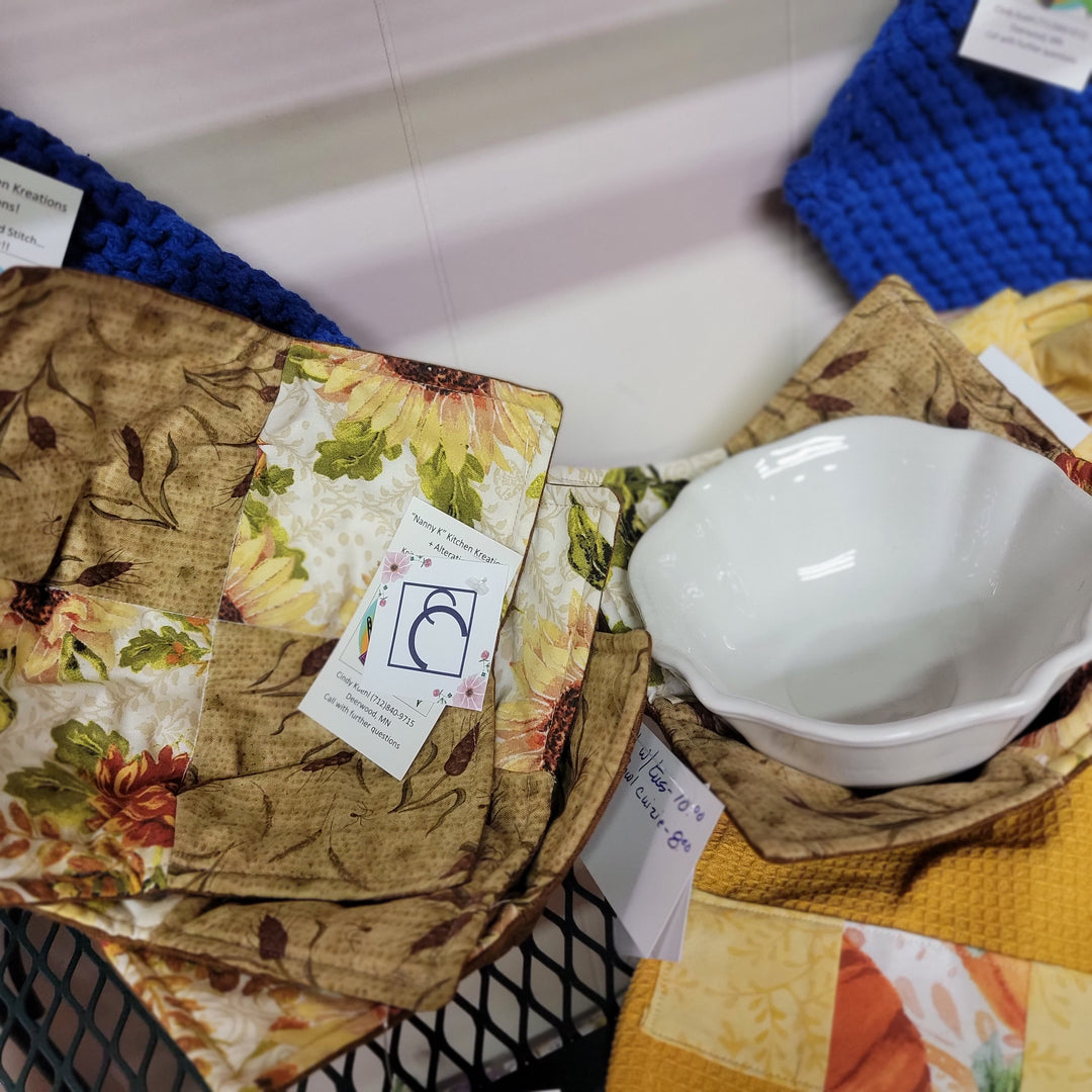 A white ceramic bowl rests on a handmade Bowl Koozie by Nanny K's Kitchen Kreations, featuring yellow floral fabric designs. The Bowl Koozies are displayed on blue and yellow textiles with tags attached to each item.