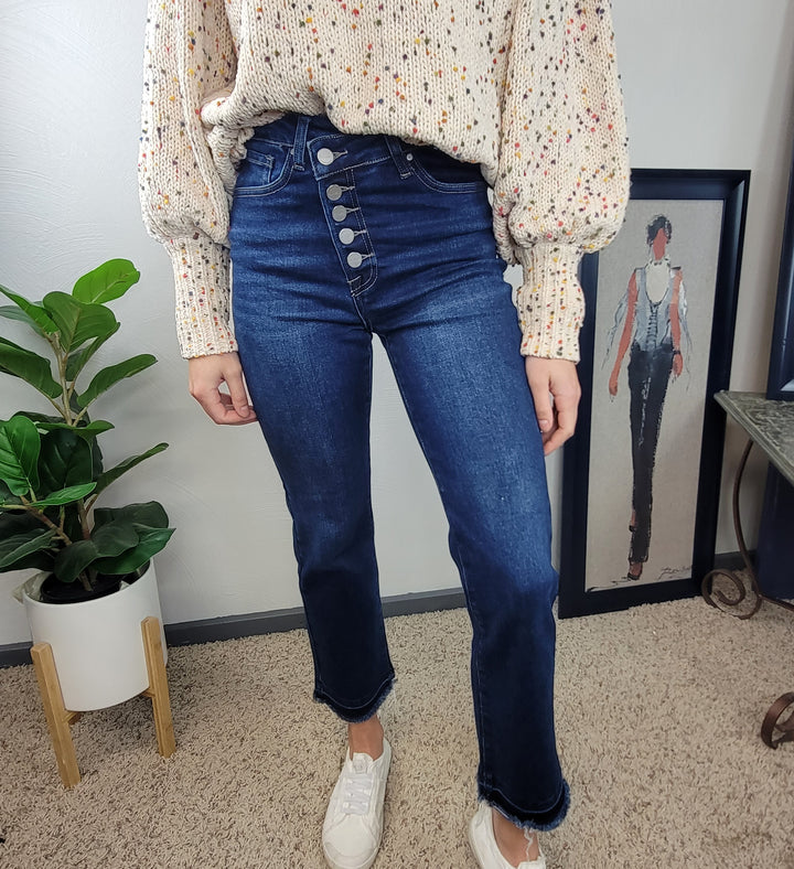 A person wearing cream, speckled sweater and Risen Haven Cross Over Straight Jeans stands indoors on a beige carpet next to a potted plant and a framed fashion illustration.