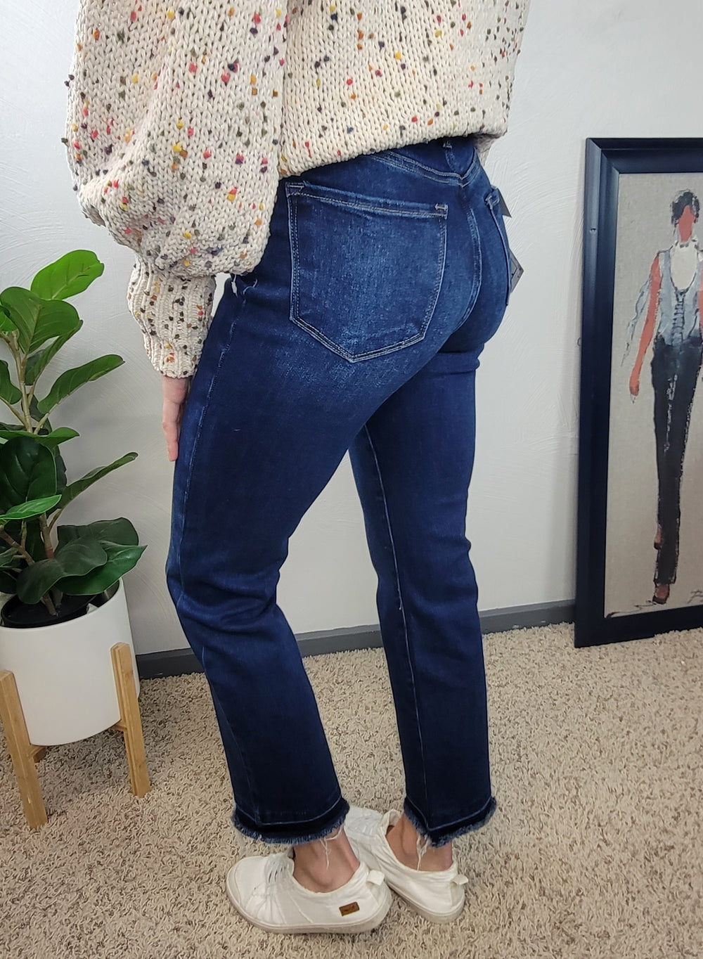 A person wearing Risen Haven Cross Over Straight Jeans, a cream sweater with colorful specks, and white shoes stands on beige carpet near a potted plant and framed artwork against a pale wall.
