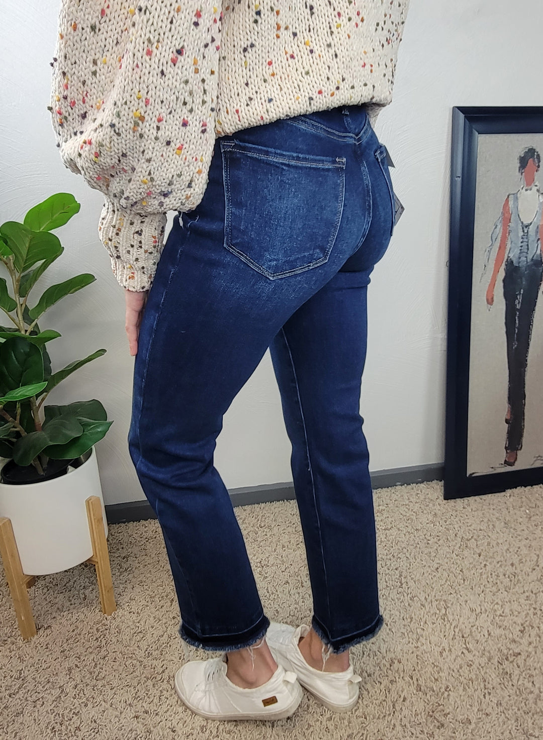 A person wearing Risen Haven Cross Over Straight Jeans, a cream sweater with colorful specks, and white shoes stands on beige carpet near a potted plant and framed artwork against a pale wall.