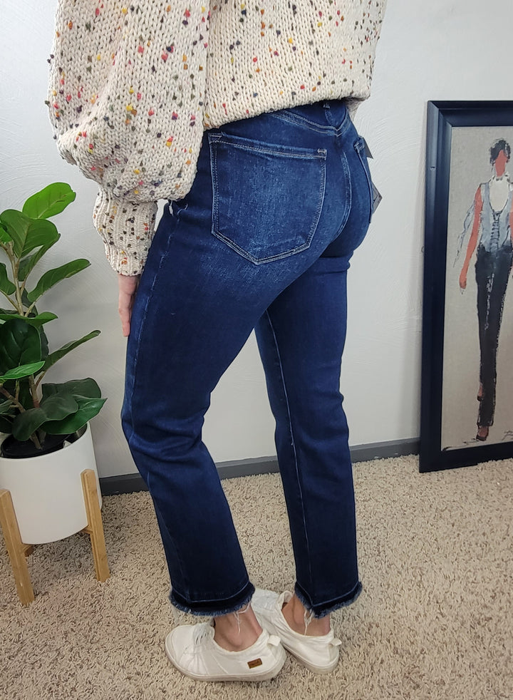 A person wearing Risen Haven Cross Over Straight Jeans, a cream sweater with colorful specks, and white shoes stands on beige carpet near a potted plant and framed artwork against a pale wall.