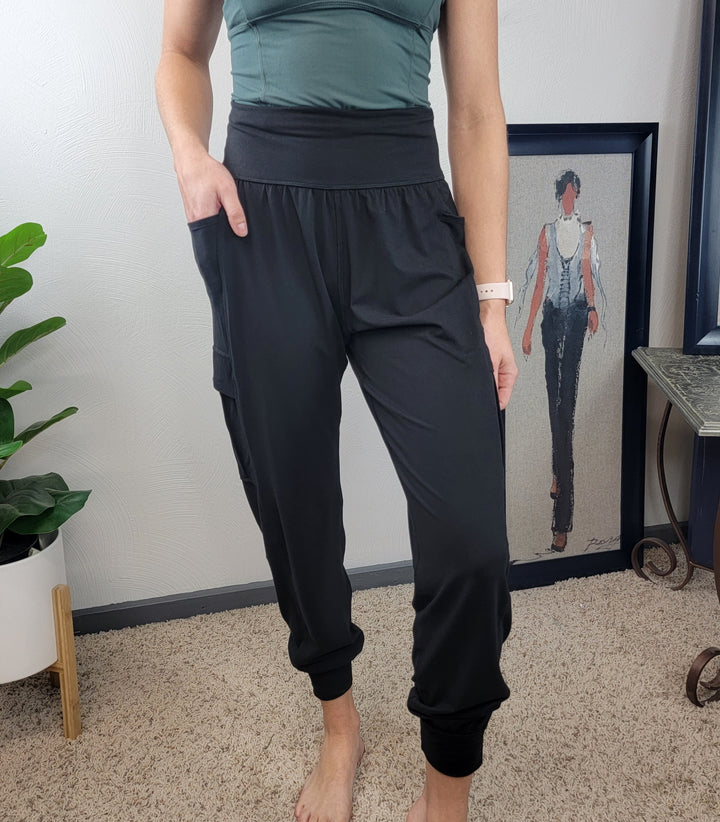 A person in Rae Mode's Carolina Joggers and a green top stands barefoot on a carpet near a potted plant, with framed artwork of a stylishly dressed woman displayed in the background.