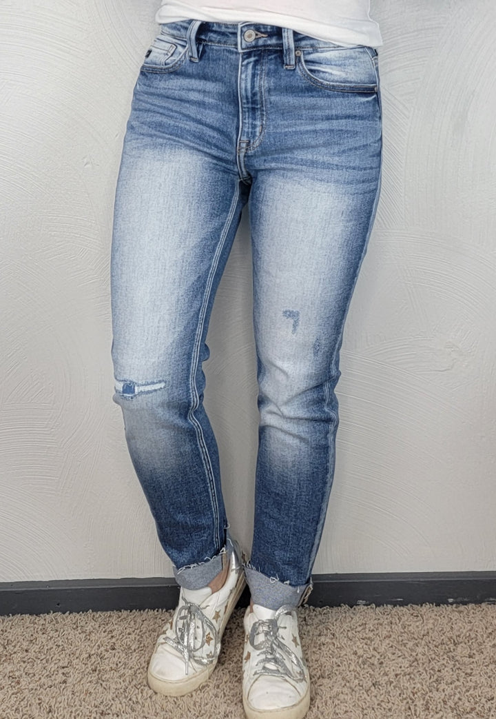 Someone wearing light blue, fitted KanCan Kaly Straight Jeans with frayed hems and white sneakers stands against a textured light gray wall on a carpeted floor.