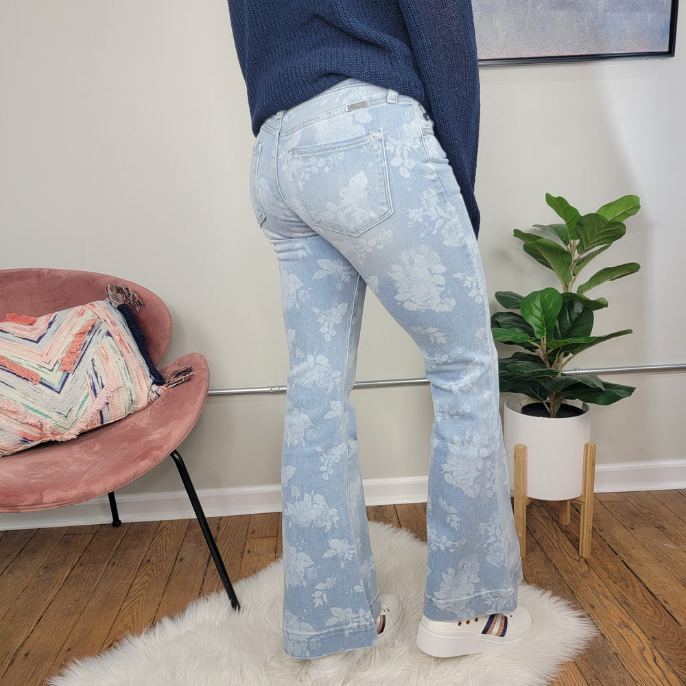 A person stands on a white rug wearing KanCan Rosa Floral Print Flares and white platform sneakers. In the background, there's a pink chair with a colorful pillow and a potted plant.