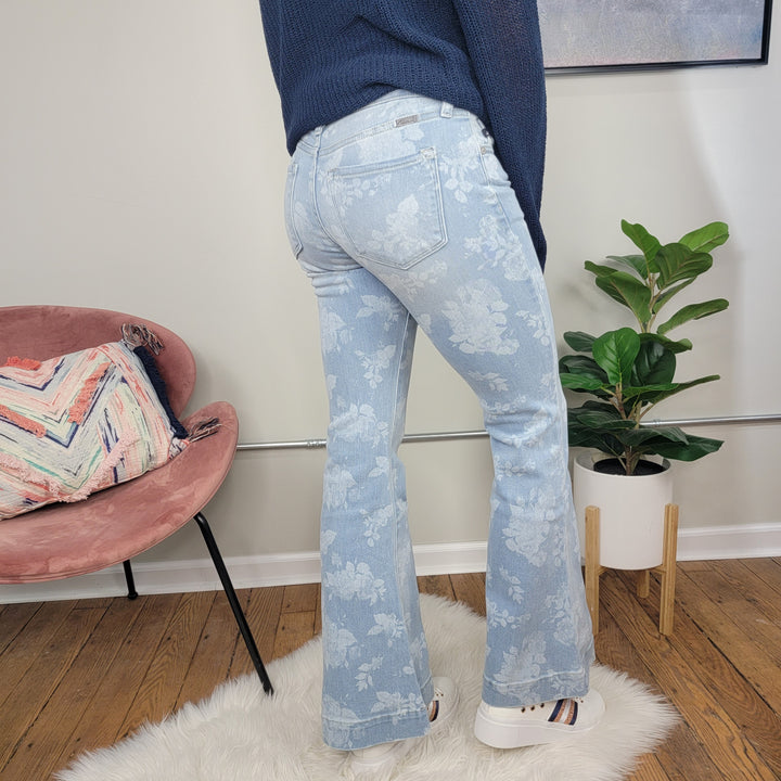 A person stands on a white rug wearing KanCan Rosa Floral Print Flares and white platform sneakers. In the background, there's a pink chair with a colorful pillow and a potted plant.