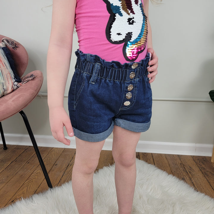 A child wearing Cutie Patootie's Kids Ava Shorts stands on a white rug in a room with wooden floors and a chair in the background.
