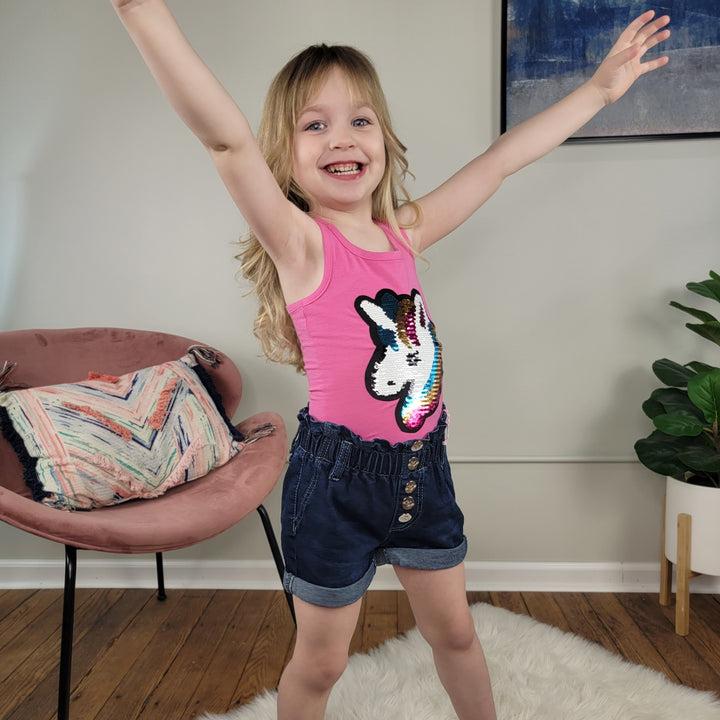 A cheerful young girl with long blond hair raises her arms in excitement, wearing a pink unicorn tank top and Cutie Patootie Kids Ava Shorts with an adjustable waist. She stands on a white rug in a stylish room with plants and a chair.