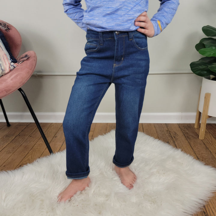 A child stands barefoot on a white fluffy rug in a living room, wearing Cutie Patootie's Kid's Jaycee Jeans—dark washed, straight-cut denim with rolled-up cuffs—and a blue long-sleeve shirt.