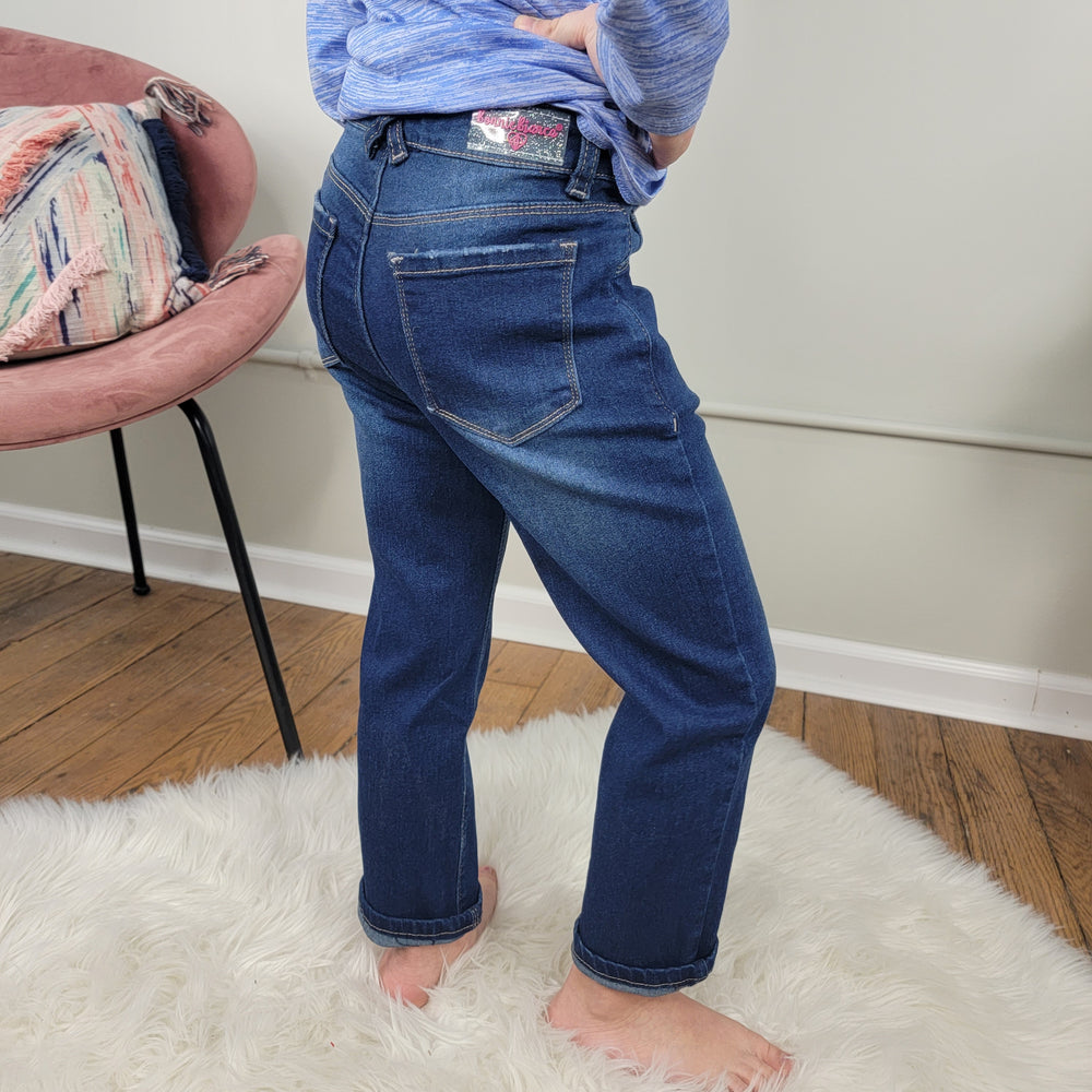 A little girl stands barefoot on a white fluffy rug next to a pink chair, wearing Cutie Patootie's Kid's Jaycee Jeans with an adjustable waist and a blue shirt, in a light room with wooden floors.