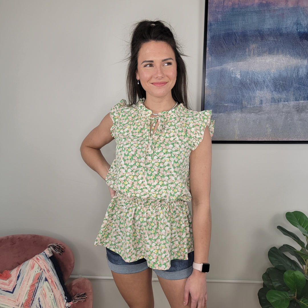 A woman in the Staccato Brie Floral Top with ruffle tie neck and denim shorts stands indoors, smiling to the side. A pink chair, green plant, and abstract wall art add charm to the scene.