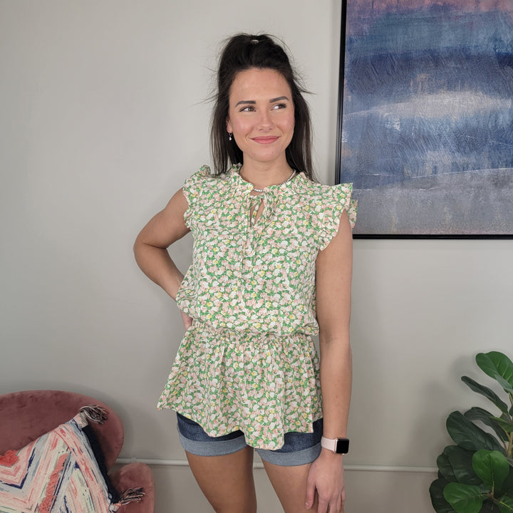 A woman in the Staccato Brie Floral Top with ruffle tie neck and denim shorts stands indoors, smiling to the side. A pink chair, green plant, and abstract wall art add charm to the scene.