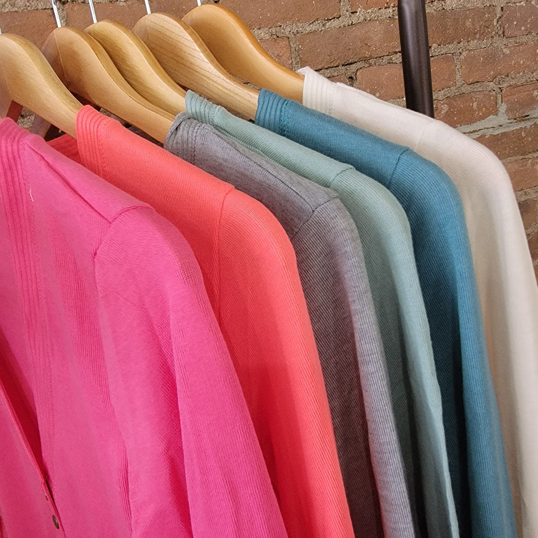 Six Zanana Long Sleeve Snap Front Cardis in pink, coral, gray, teal, light blue, and off-white hang on wooden hangers against a brick wall. Perfect for layering, these essentials suit any season.