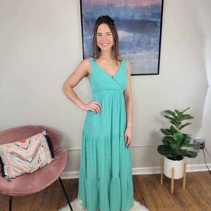 A smiling woman with long brown hair wears the Timing Vivian Strappy Maxi Dress and stands indoors by a pink chair with a pillow, a potted plant, and abstract wall art in the background.