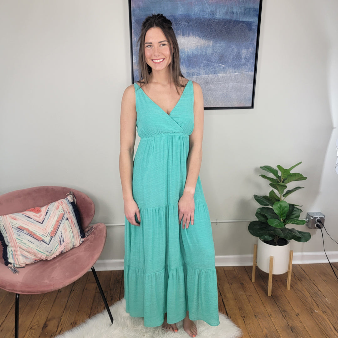 A woman wears the Timing Vivian Strappy Maxi Dress, a sleeveless jade maxi, smiling in a room with wooden floors. Behind her are a pink chair with patterned pillow, potted plant, and abstract painting—perfect as a summer wedding dress.