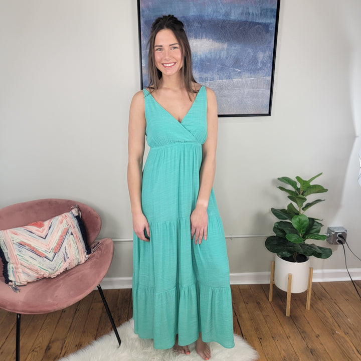 A woman wears the Timing Vivian Strappy Maxi Dress, a sleeveless jade maxi, smiling in a room with wooden floors. Behind her are a pink chair with patterned pillow, potted plant, and abstract painting—perfect as a summer wedding dress.