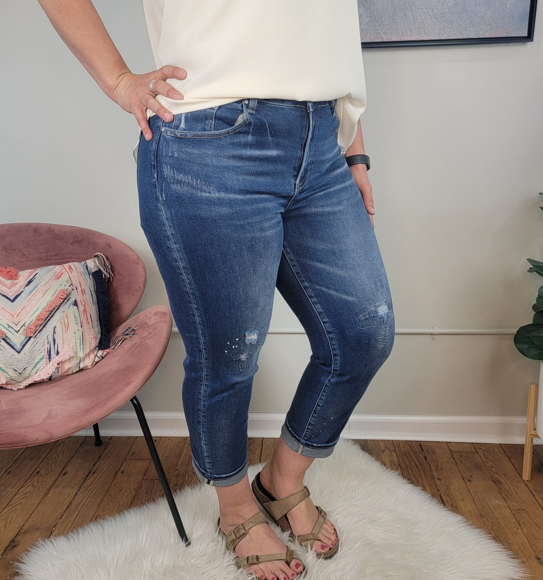 A person wears Risen Valorie Jeans, a white top, and beige sandals while standing on a white rug. In the background are a pink chair with a patterned pillow and a green plant.