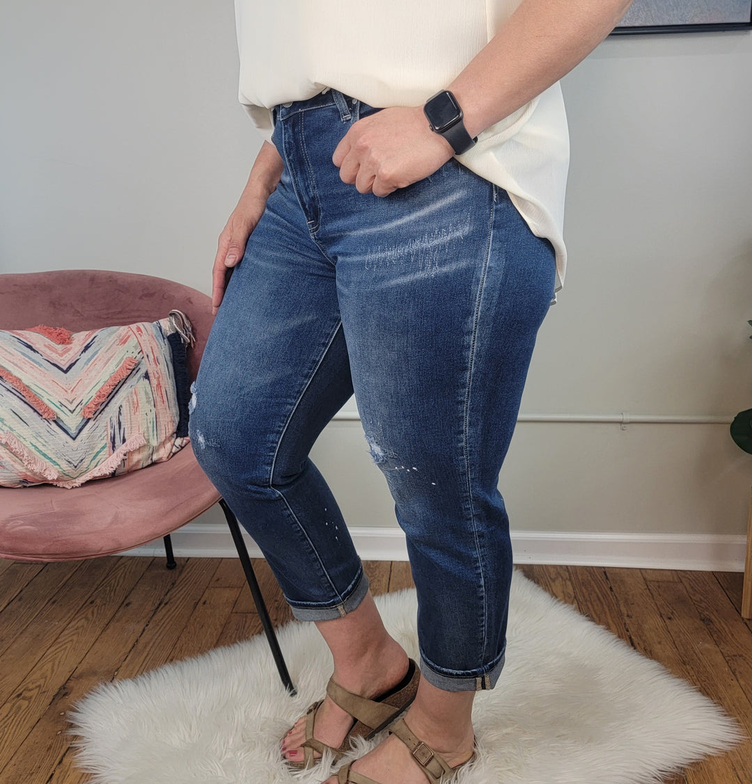 Someone stands on a white rug wearing a white top, beige sandals, a smartwatch, and Risen Valorie Jeans in dark wash. In the background is a pink chair with a patterned pillow on a wooden floor.